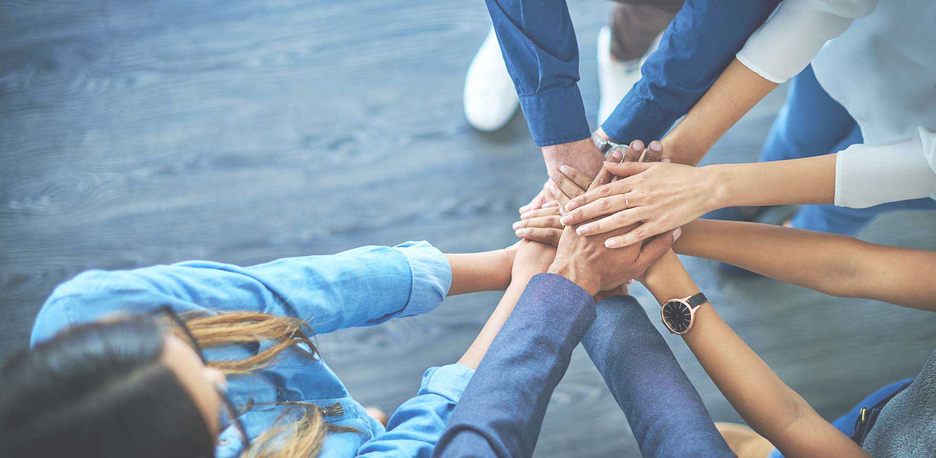 Mehrere Hände stapeln sich in der Mitte, während Menschen zusammenarbeiten. Der Boden ist aus Holz und die Atmosphäre ist kooperativ.