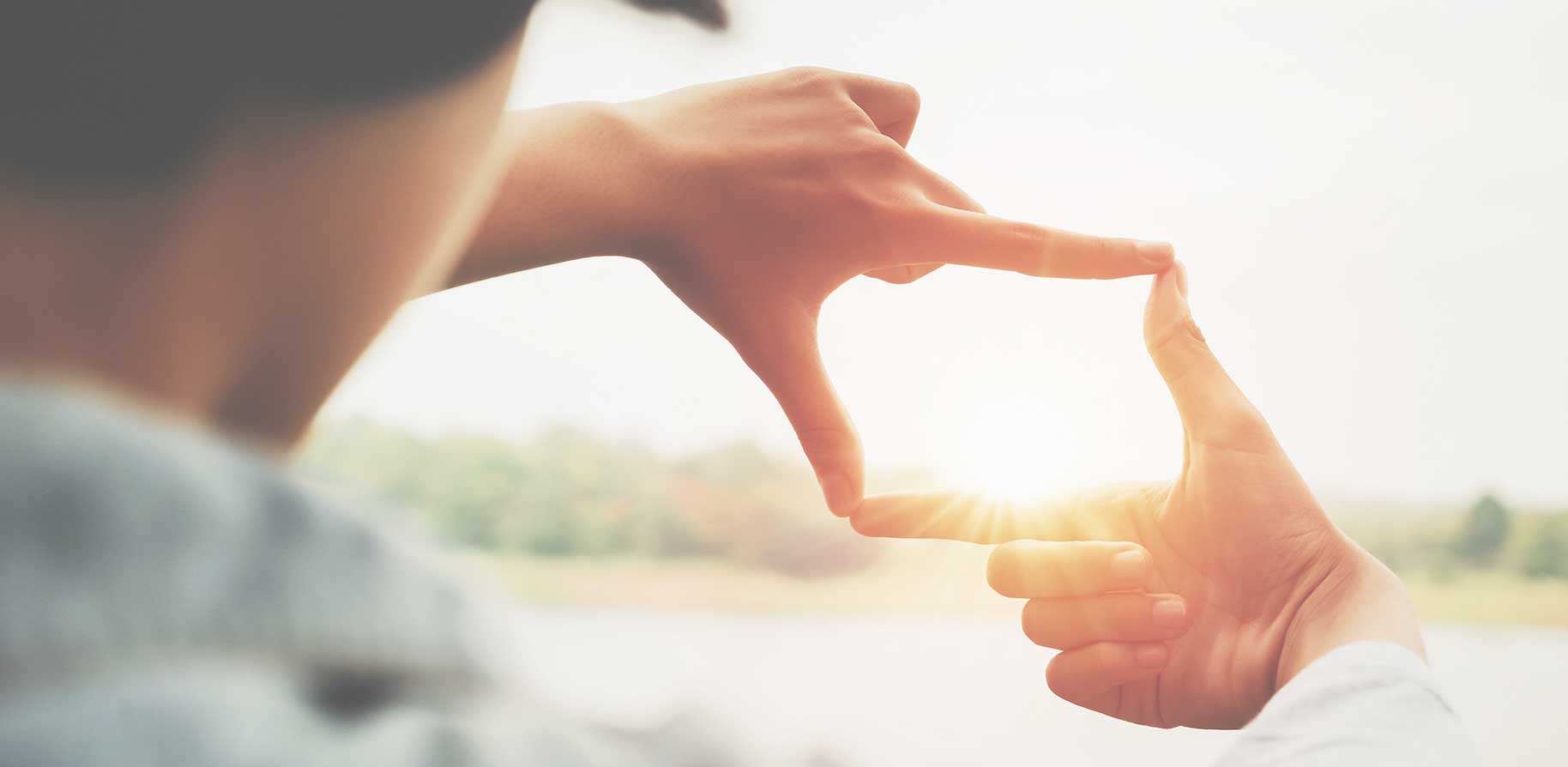 Eine Person formt mit ihren Händen ein Rechteck, durch das die Sonne scheint, während im Hintergrund eine unscharfe Landschaft sichtbar ist.