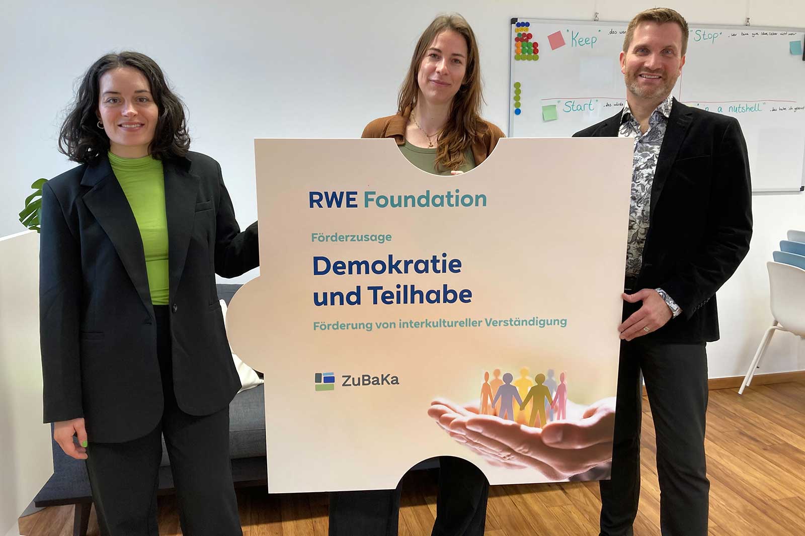 Drei Personen halten ein großes Schild, das die RWE Stiftung und den Text 'Demokratie und Teilhabe' zeigt.