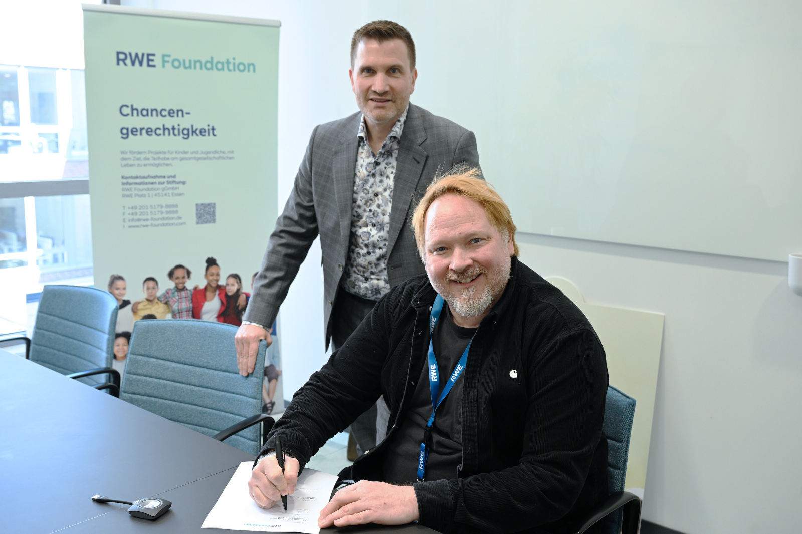 A man in a black shirt is signing a document at a conference table with an RWE Foundation poster in the background.