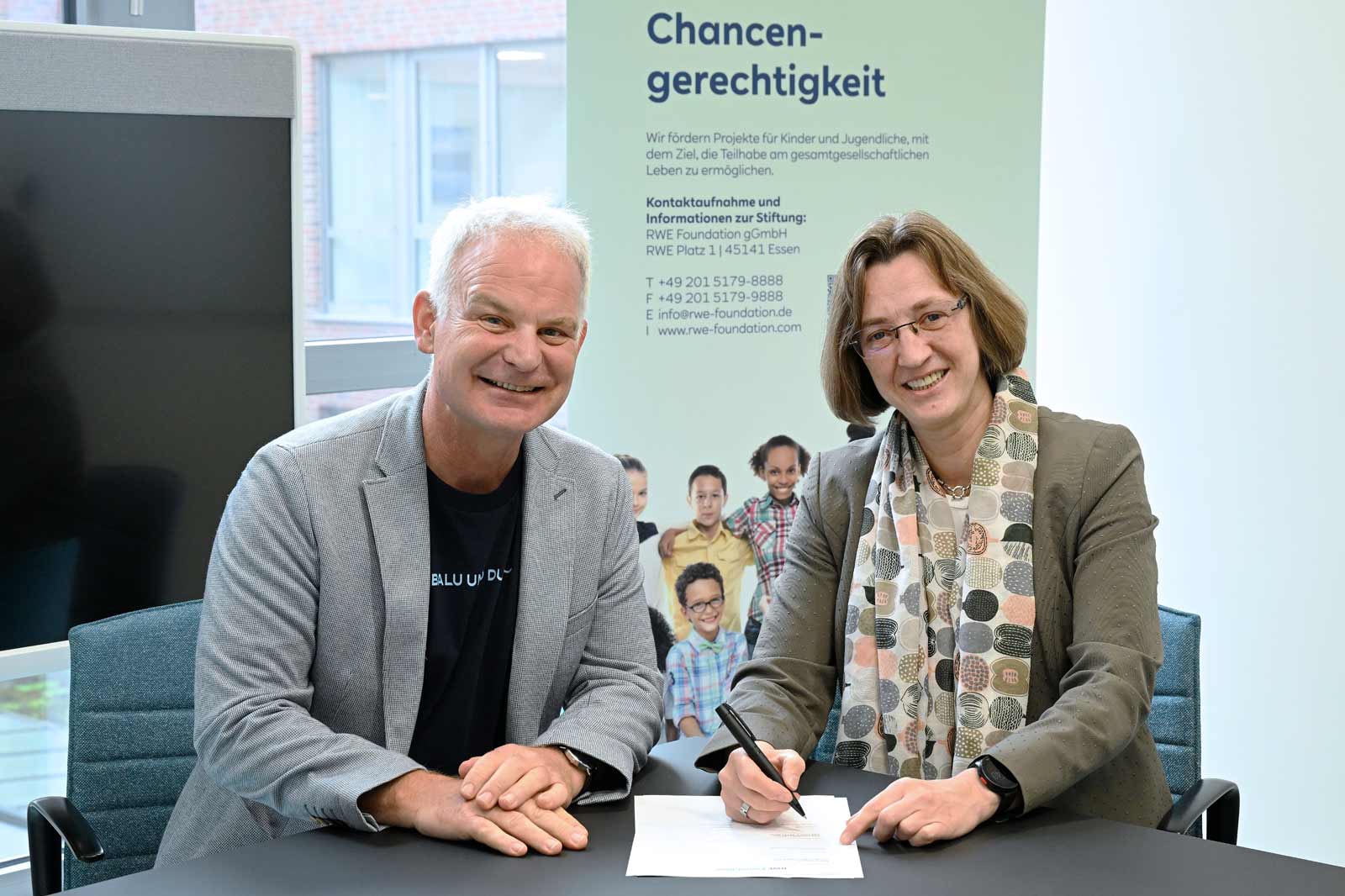 Two individuals are signing a contract at a table, with a poster about equal opportunities in the background.