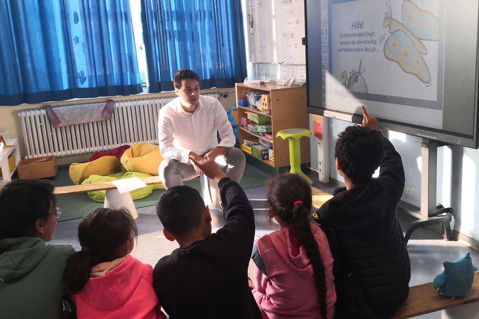 Eine Gruppe von Kindern sitzt in einer Lernumgebung und hört einem Lehrer zu, während sie auf einen Bildschirm zeigen.