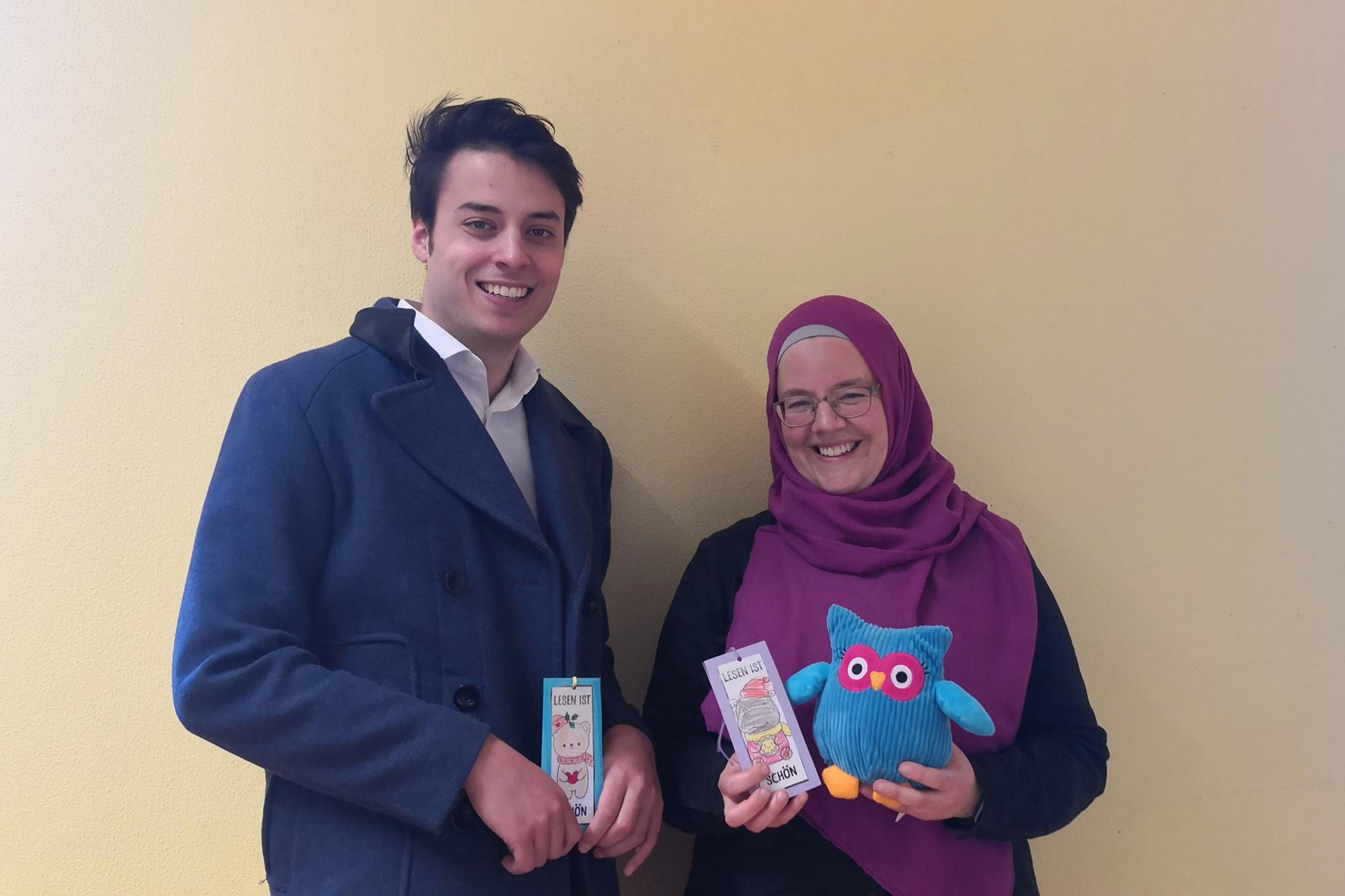 Zwei Personen halten Bücher und ein Plüschtier vor einer gelben Wand.