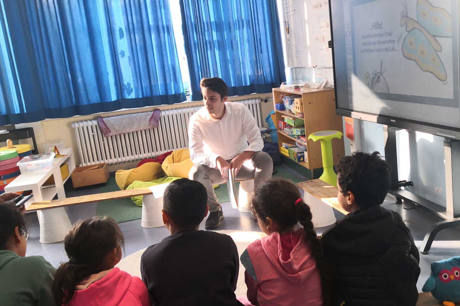 Ein Lehrer sitzt vor einer Gruppe von Kindern in einem Klassenzimmer mit bunten Kissen und einem interaktiven Bildschirm.