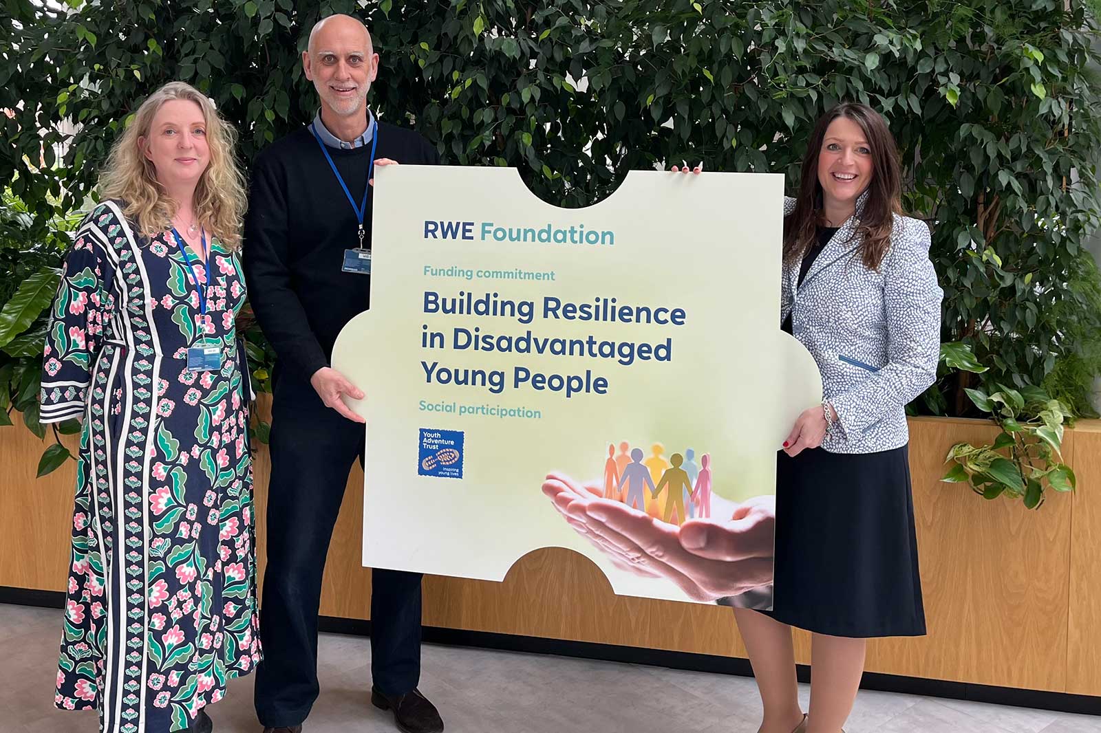 Drei Personen halten ein Schild mit der Aufschrift 'Building Resilience in Disadvantaged Young People' vor einer grünen Pflanzenwand.