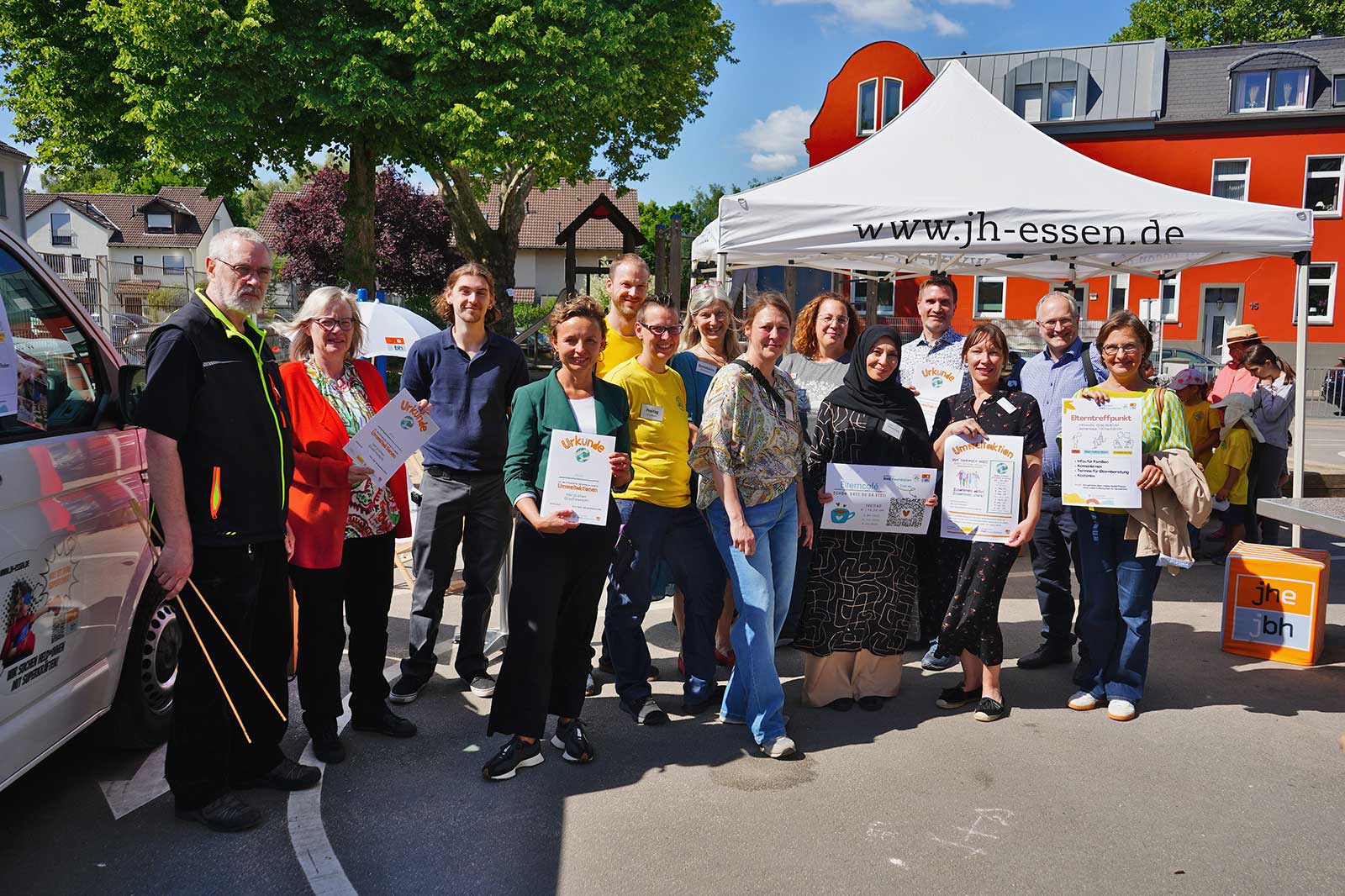 A diverse group of people holding certificates stands under a canopy, with a festive atmosphere in a sunny outdoor setting.