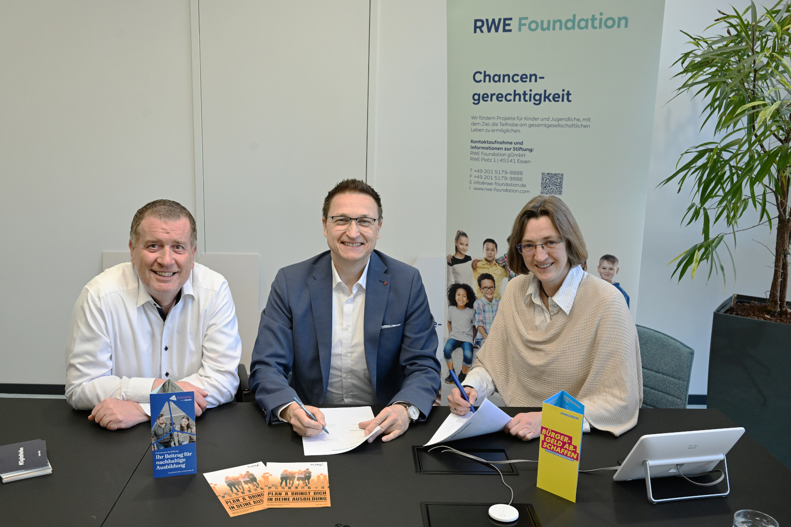 Three individuals sit at a table with documents and brochures, with the RWE Foundation logo visible in the background.