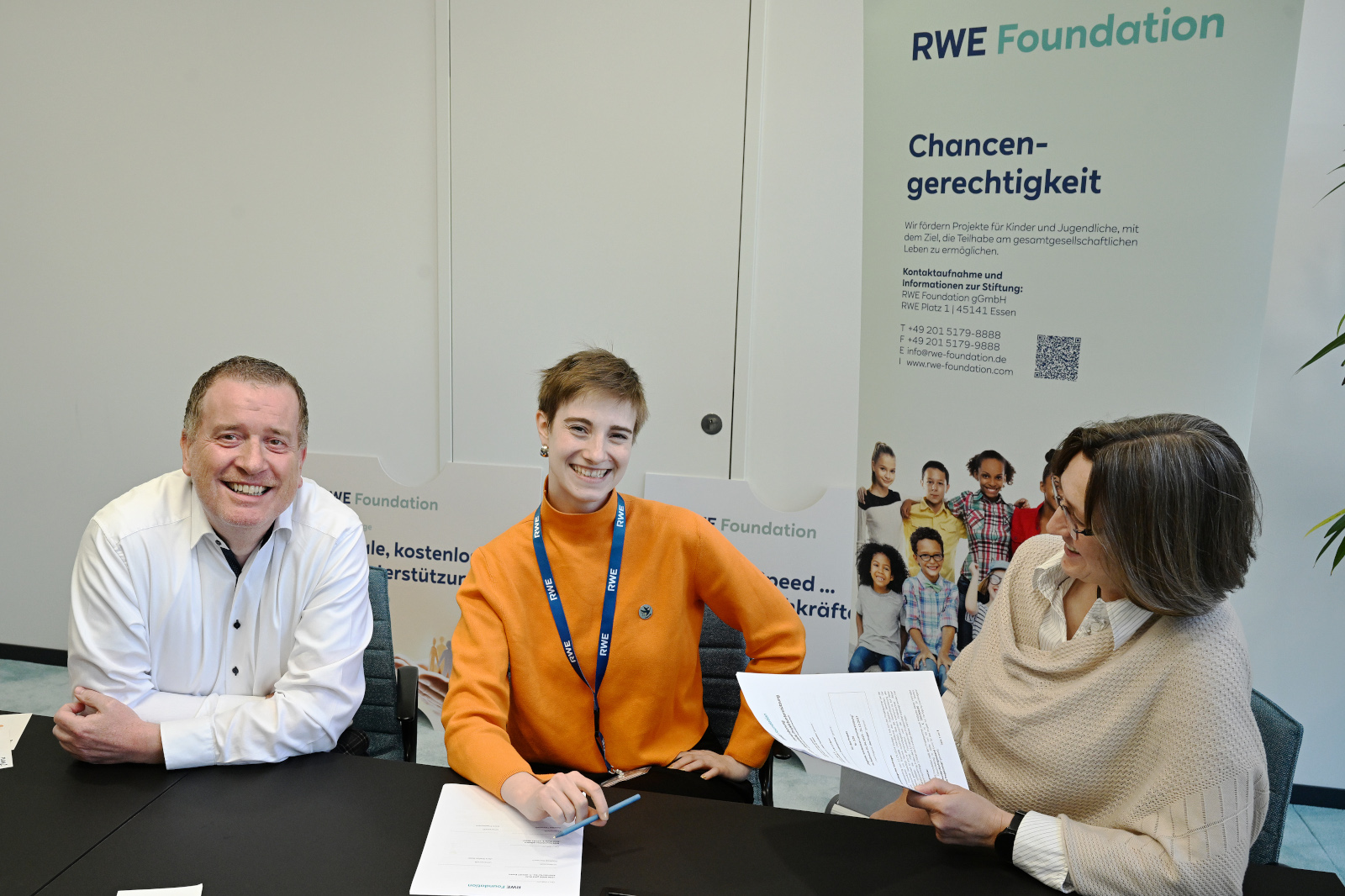 Drei Personen an einem Tisch in einem Besprechungsraum, eine Person trägt ein oranges Oberteil.