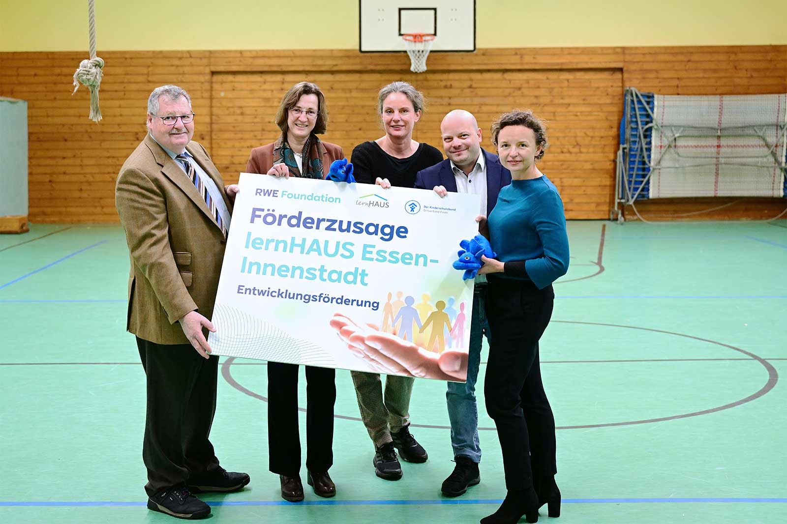 Eine Gruppe von Menschen hält ein großes Plakat mit der Aufschrift 'Förderzusage lernHAUS Essen-Innenstadt' in einer Sporthalle.