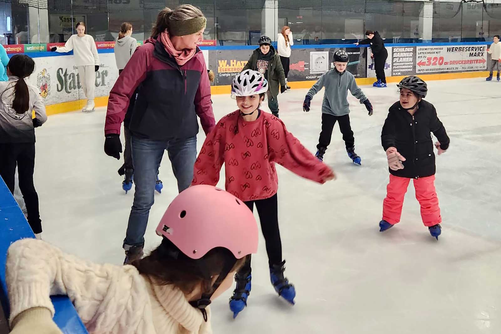 Children ice skating in a rink, supervised by an adult, with some wearing helmets and colourful clothing.