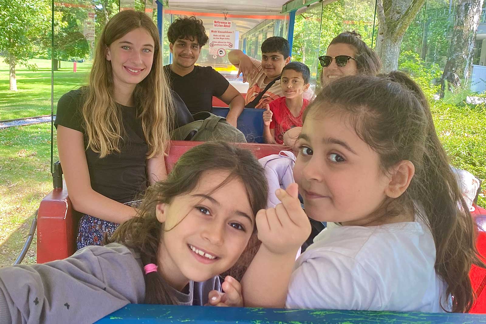Gruppe von Kindern und Jugendlichen auf einem Zug im Freien, umgeben von Bäumen und Grünflächen.