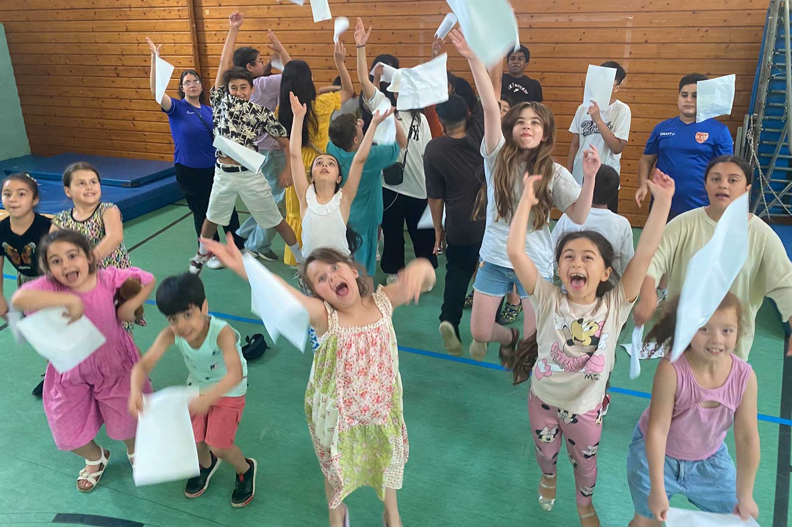 Eine lebhafte Gruppe von Kindern springt fröhlich und wedelt mit weißen Blättern in einer Turnhalle.