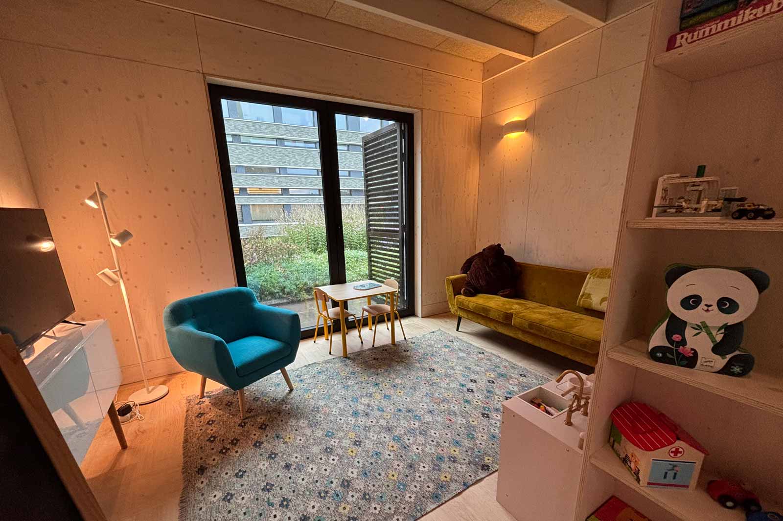 A cosy room featuring a yellow sofa, a blue chair, and a small table. There is a window overlooking the garden.