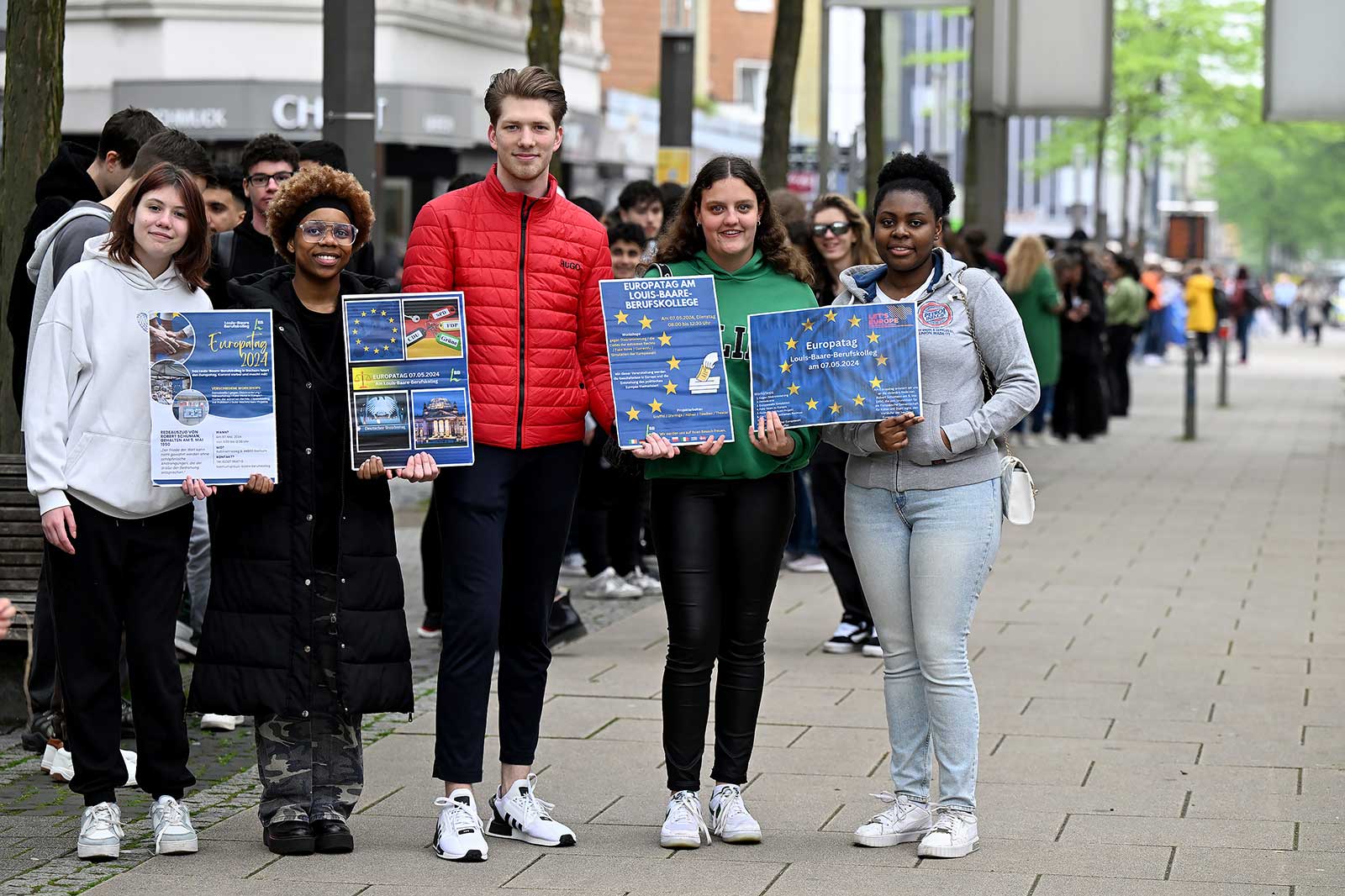Groups of people holding posters for Europe Day stand on a street, surrounded by trees.