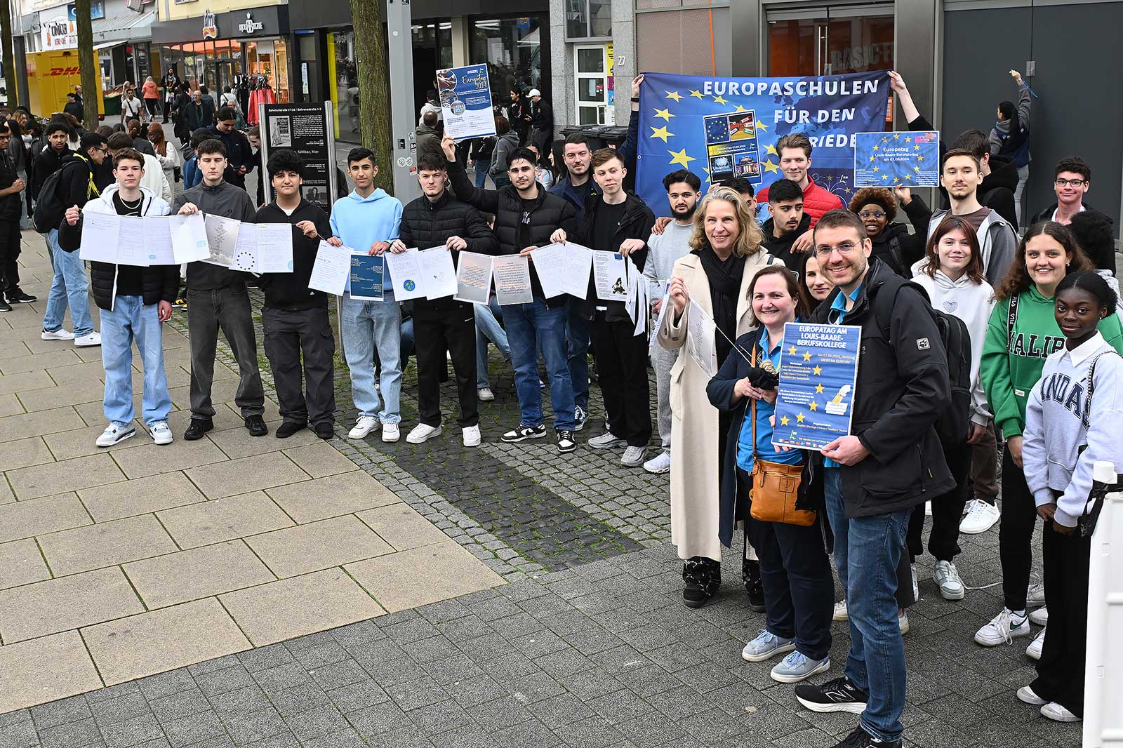 Eine Gruppe von jungen Menschen hält Plakate und Banner mit dem Text 'EUROPASCHULEN FÜR DEN FRIEDEN' auf einem Platz.