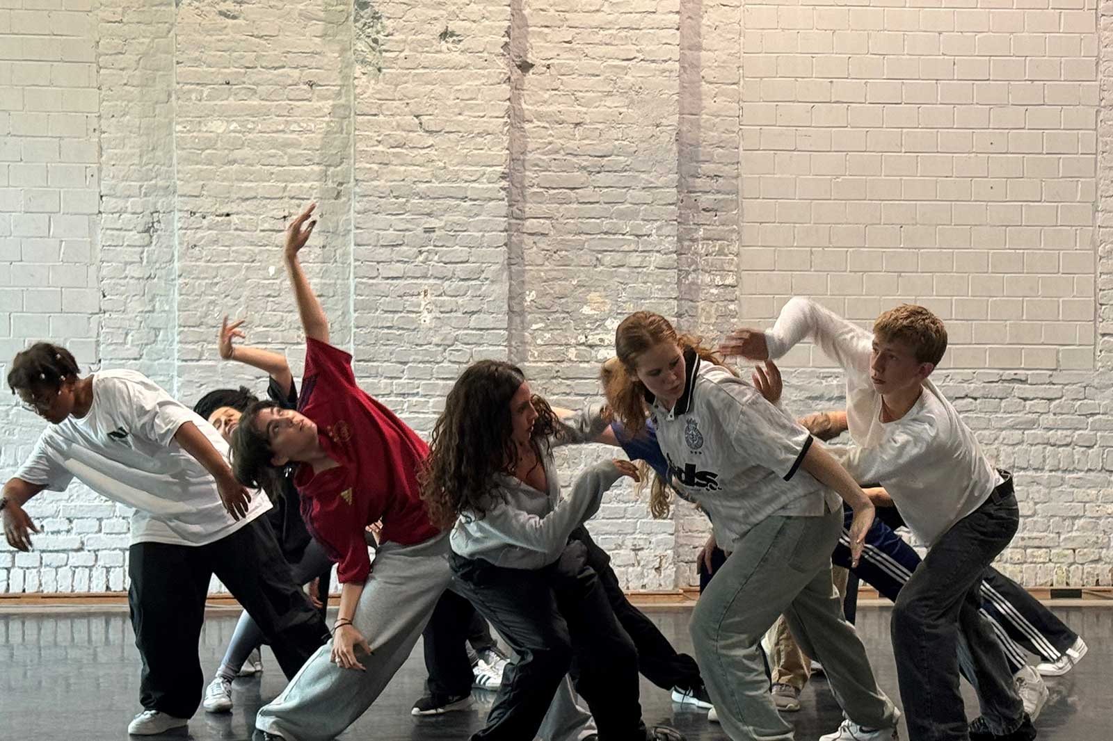 Eine Gruppe von Tänzern führt einen synchronen Tanz in einem Studio auf. Der Hintergrund besteht aus weißen Ziegeln.