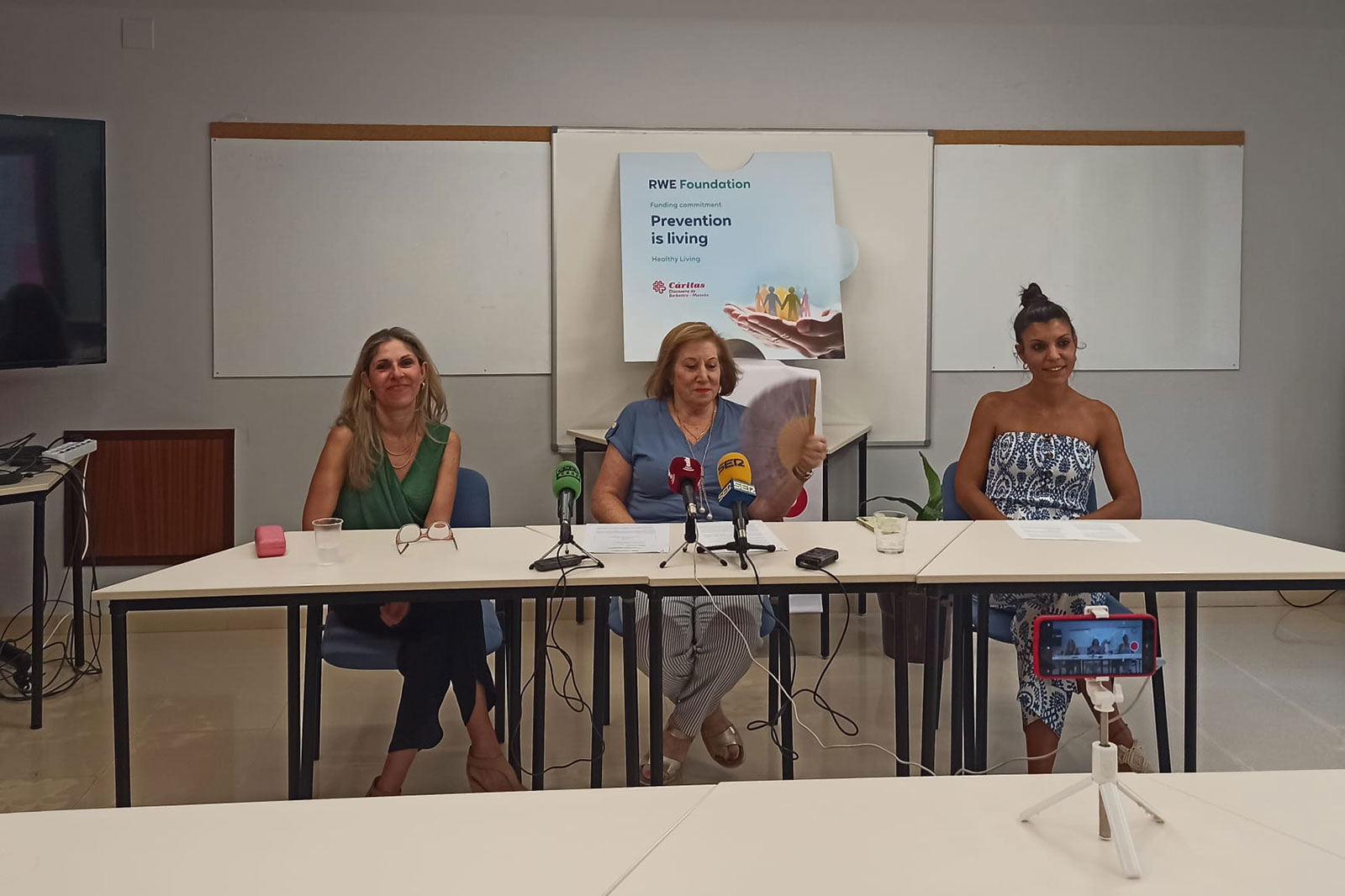Three women participate in a press conference with a poster stating 'Prevention is living' in the background.