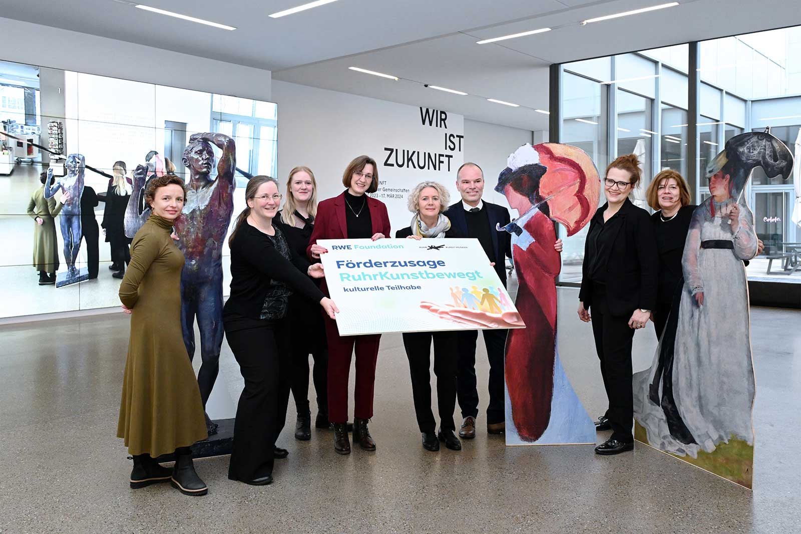 A group of people holds a sign reading 'Förderzusage RuhrKunstbewegt' in a modern gallery space.