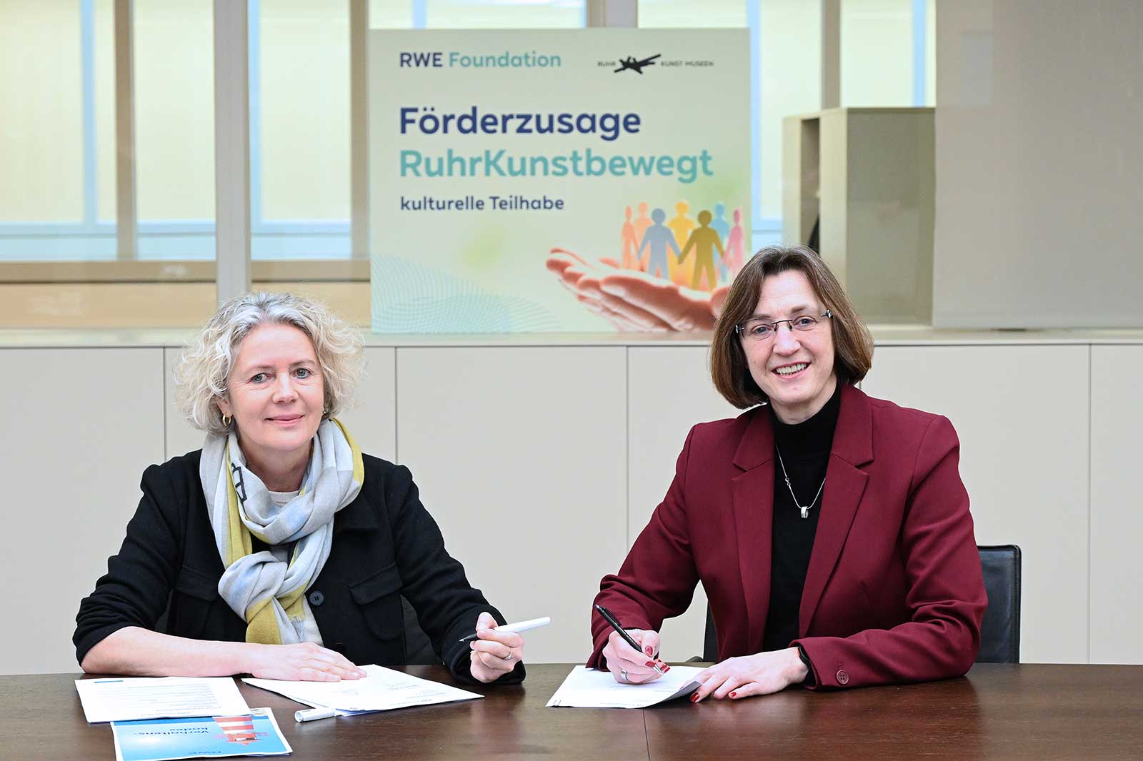 Two women are signing documents. A banner in the background reads 'Förderzusage RuhrKunstbewegt'.