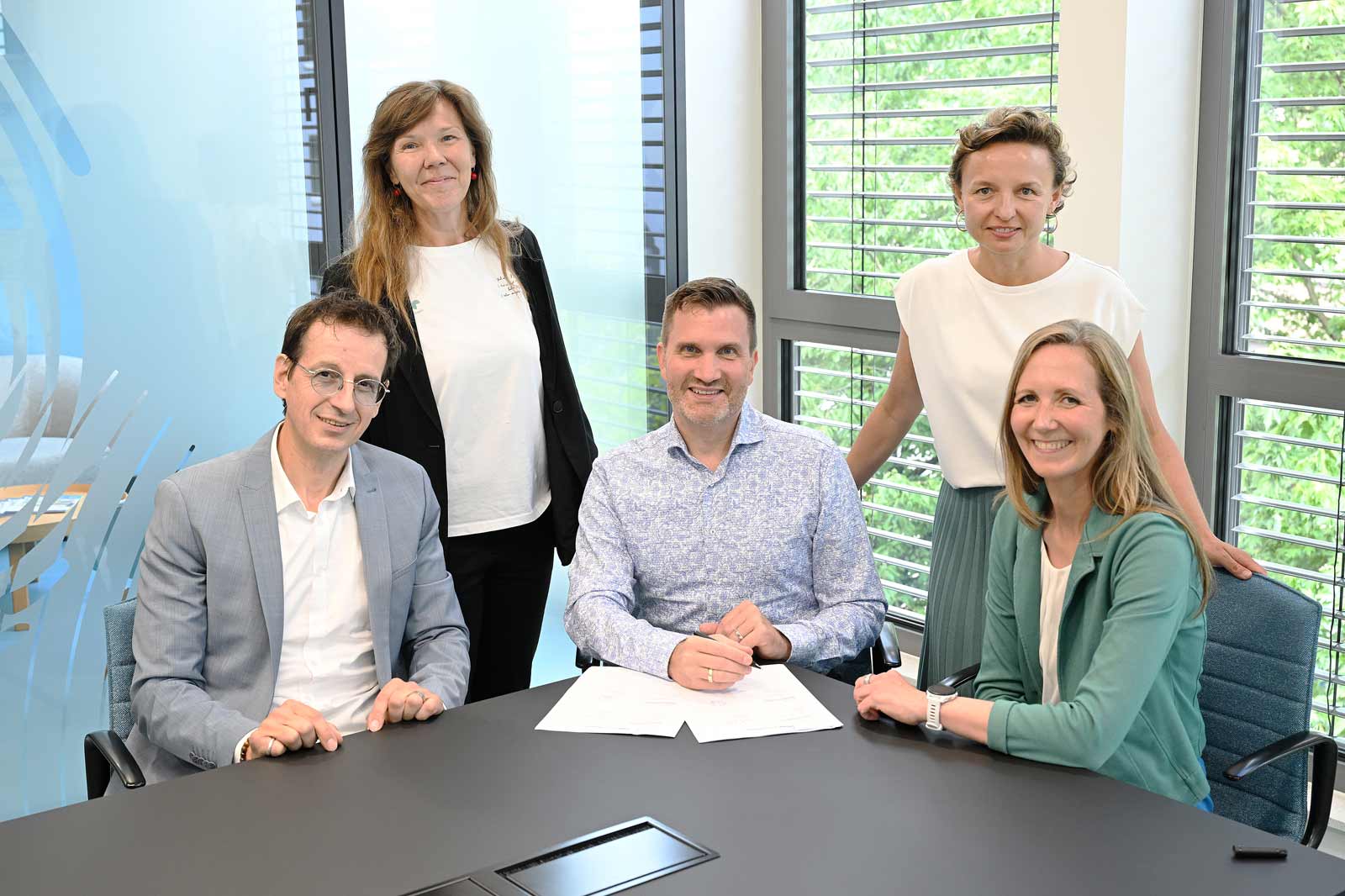 A group of five people stands around a table with documents on it, posing in a modern office.