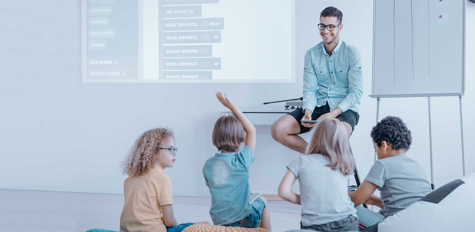 A teacher sits on a platform while children engage in a coding lesson. A projection screen displays programming commands.