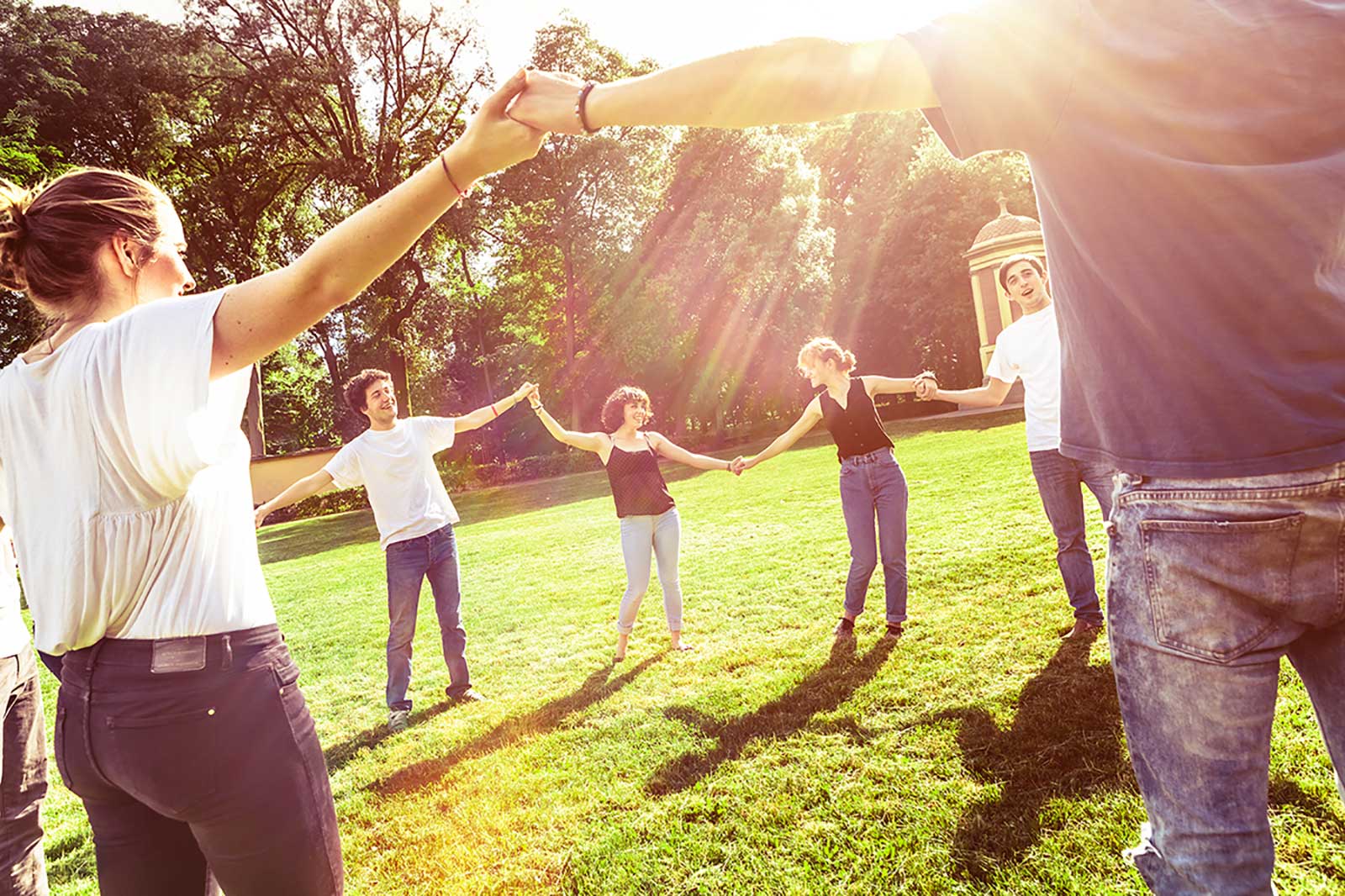 A group of people forms a circle in the park, holding hands as the sun shines behind them.