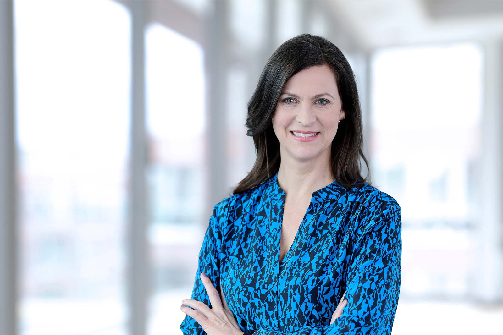 Eine Frau trägt ein blaues Musterhemd und steht mit verschränkten Armen in einem modernen Raum mit viel Tageslicht. Portrait von Birgit Hiller.