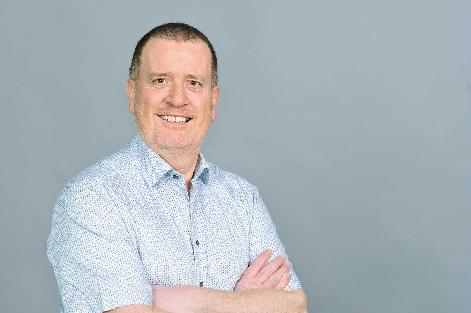 A man wearing a patterned shirt stands with his arms crossed against a grey background.