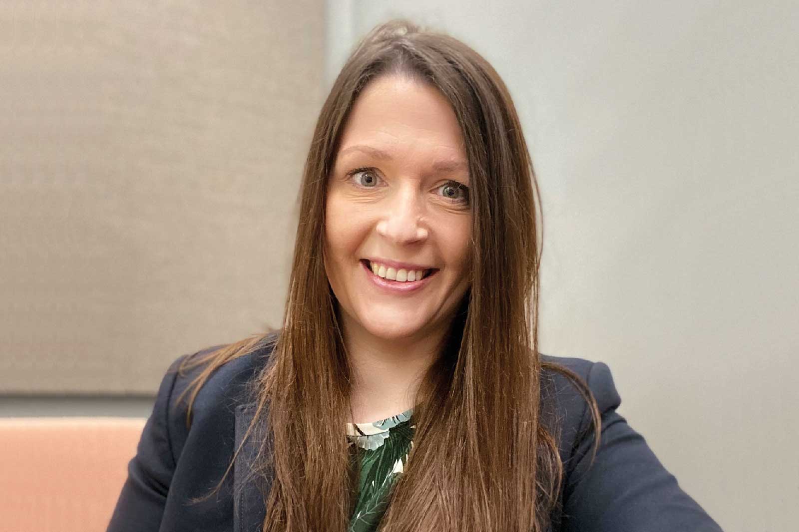 A person with long brown hair is wearing a black blazer and a green top with a leaf pattern.