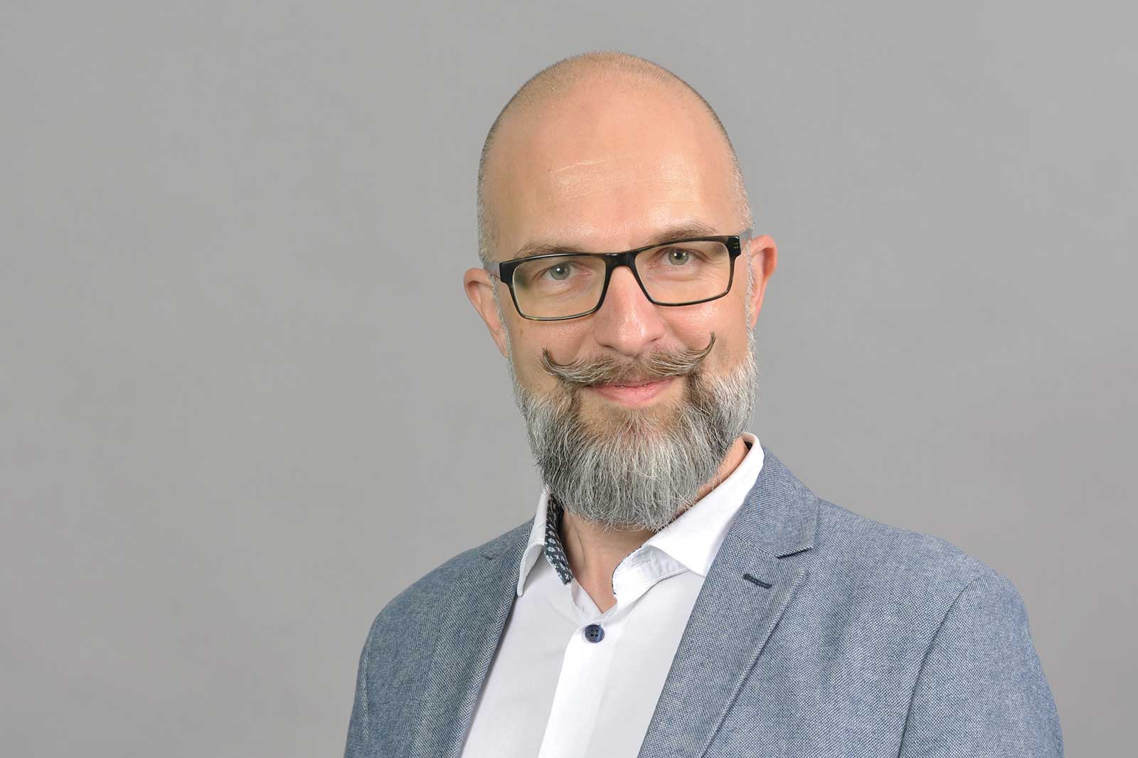 A man in a grey suit with a beard stands against a grey background.