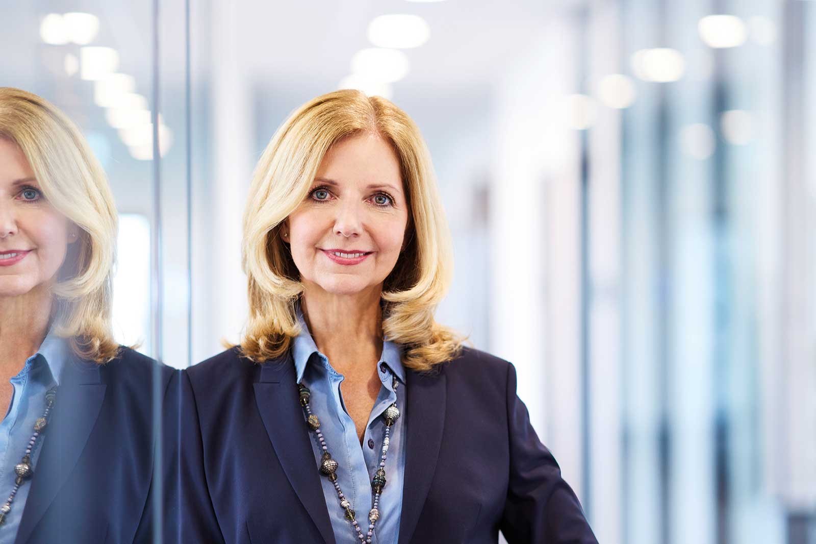 A blonde woman in a blazer stands in front of a mirror in a modern office with bright lights.