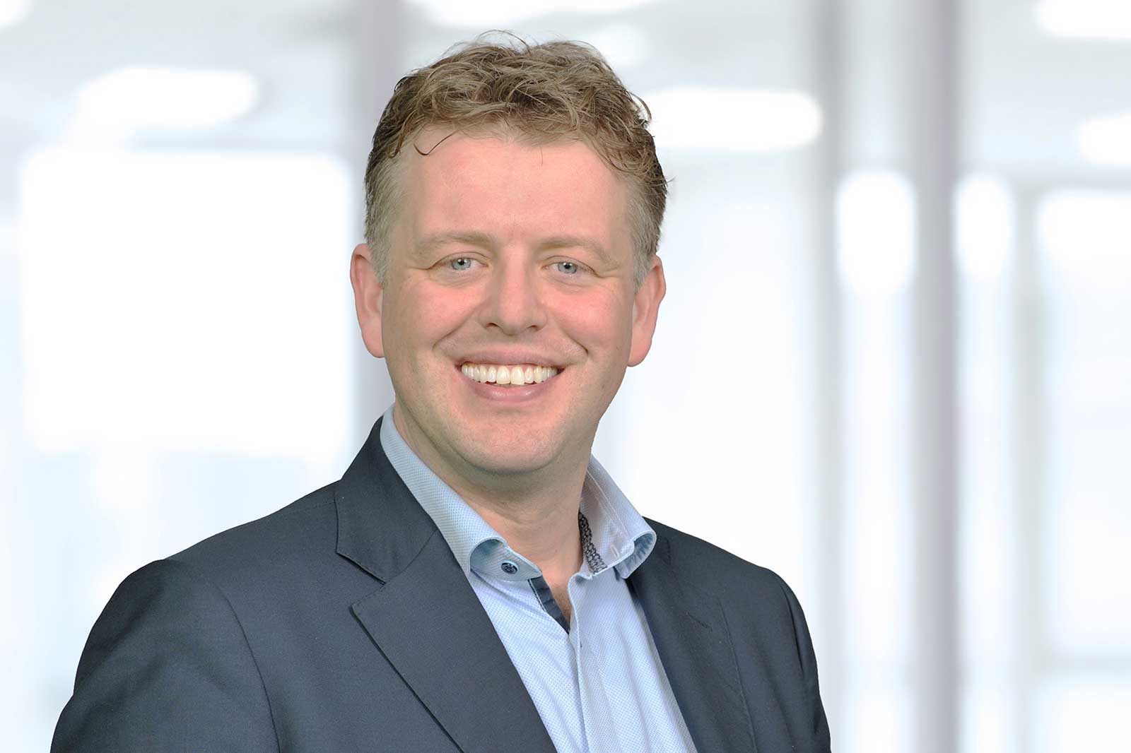 A man in a suit with a light blue shirt stands against a blurred office background.