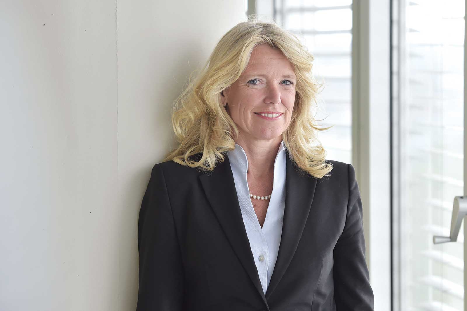 A woman with long, wavy blonde hair wearing a black blazer and a light blue shirt, standing next to a white wall.