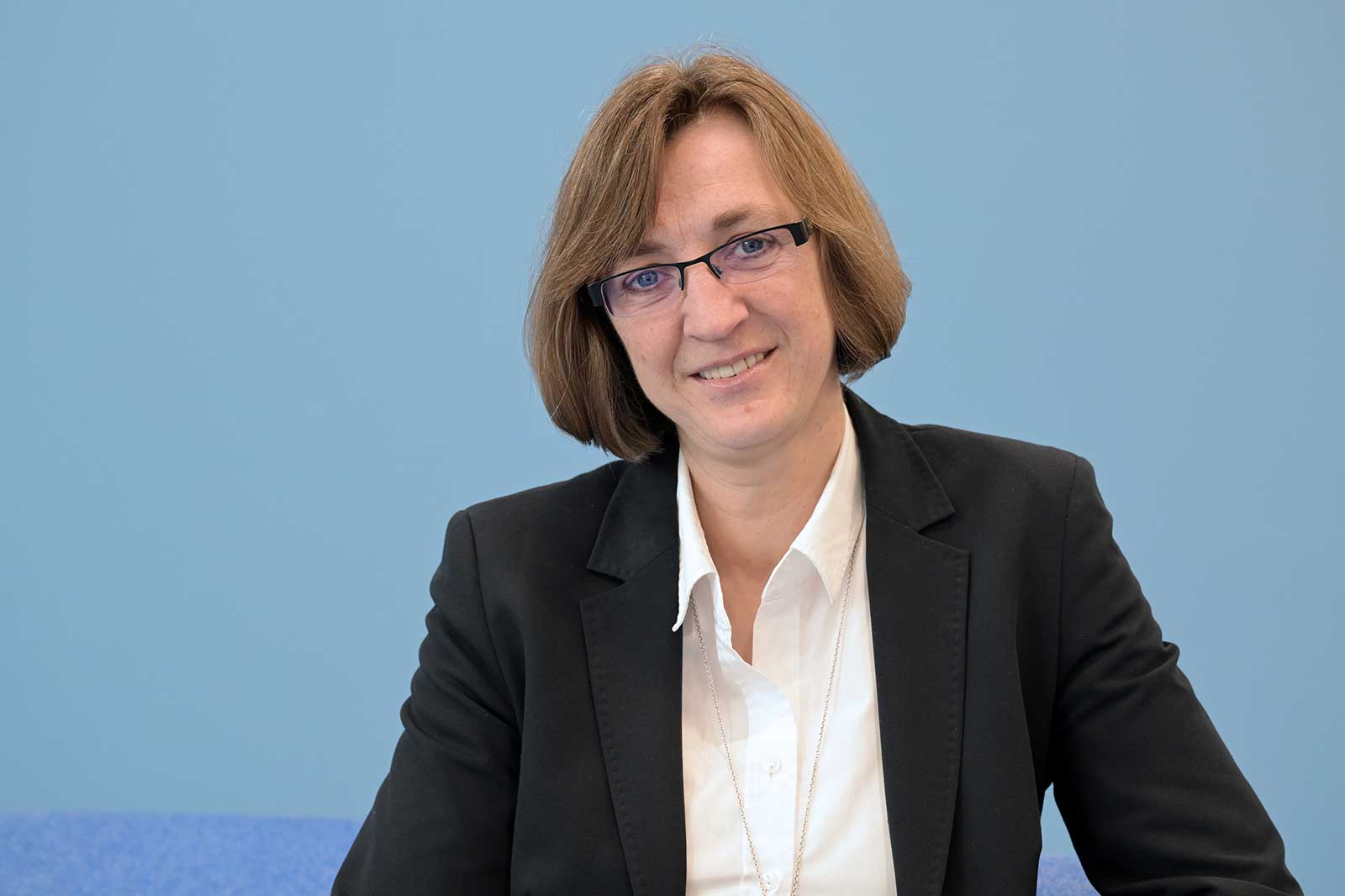 A person in formal attire sits against a light blue background. She has shoulder-length brown hair.