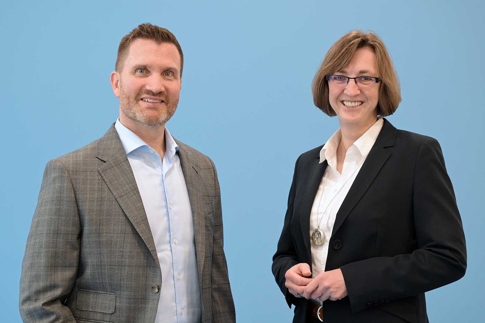 Ein Mann in einem grauen Anzug und eine Frau im schwarzen Anzug stehen vor einem hellblauen Hintergrund, beide tragen formelle Kleidung.