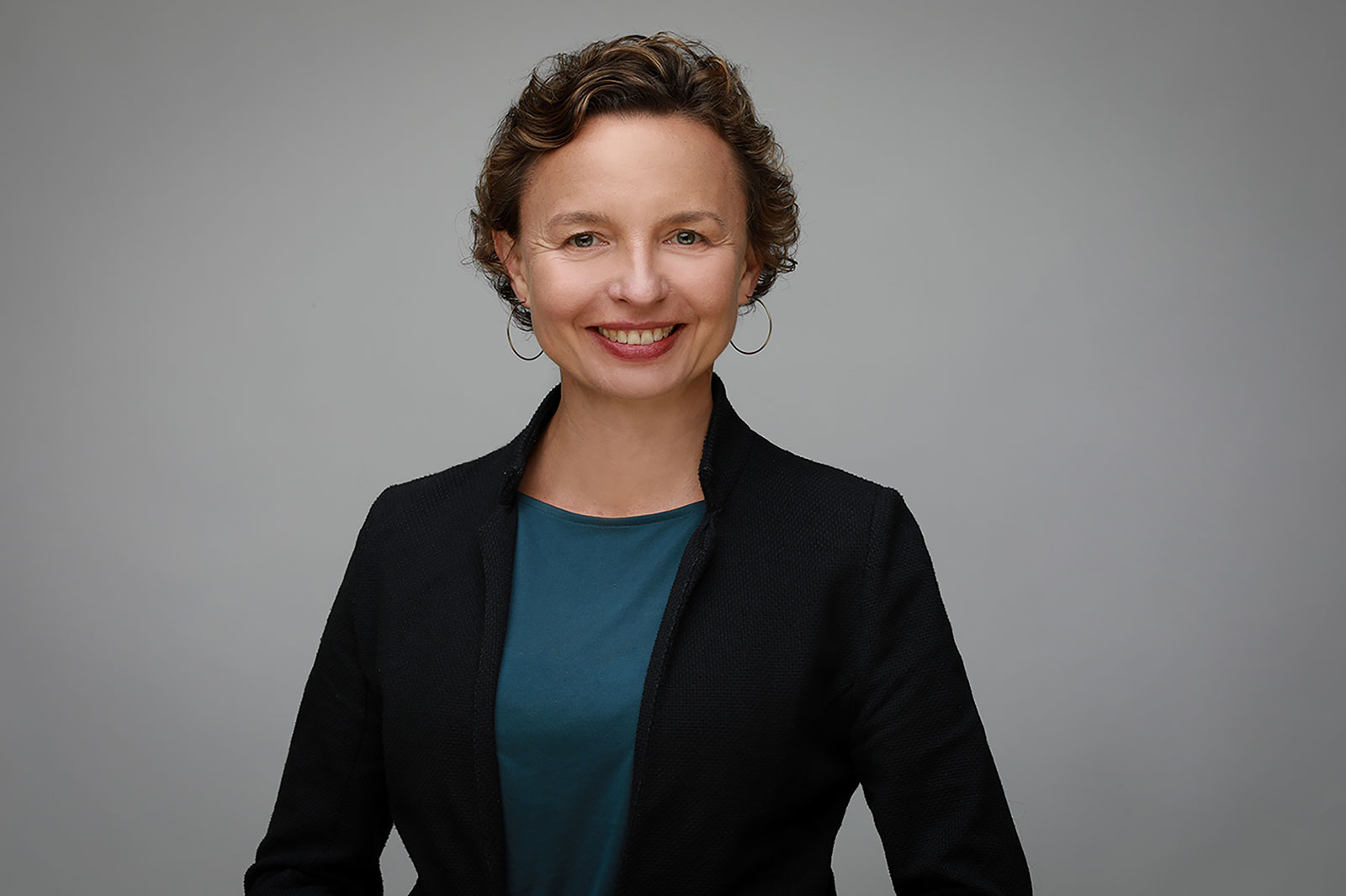 Die Person trägt eine schwarze Jacke und ein blaues T-Shirt. Der Hintergrund ist grau.