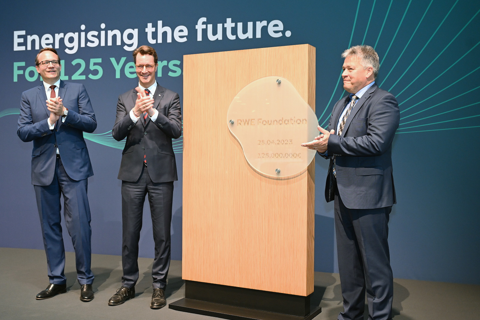 Three men applaud in front of a wooden plaque inscribed with 'RWE Foundation' and the date 25.04.2023.