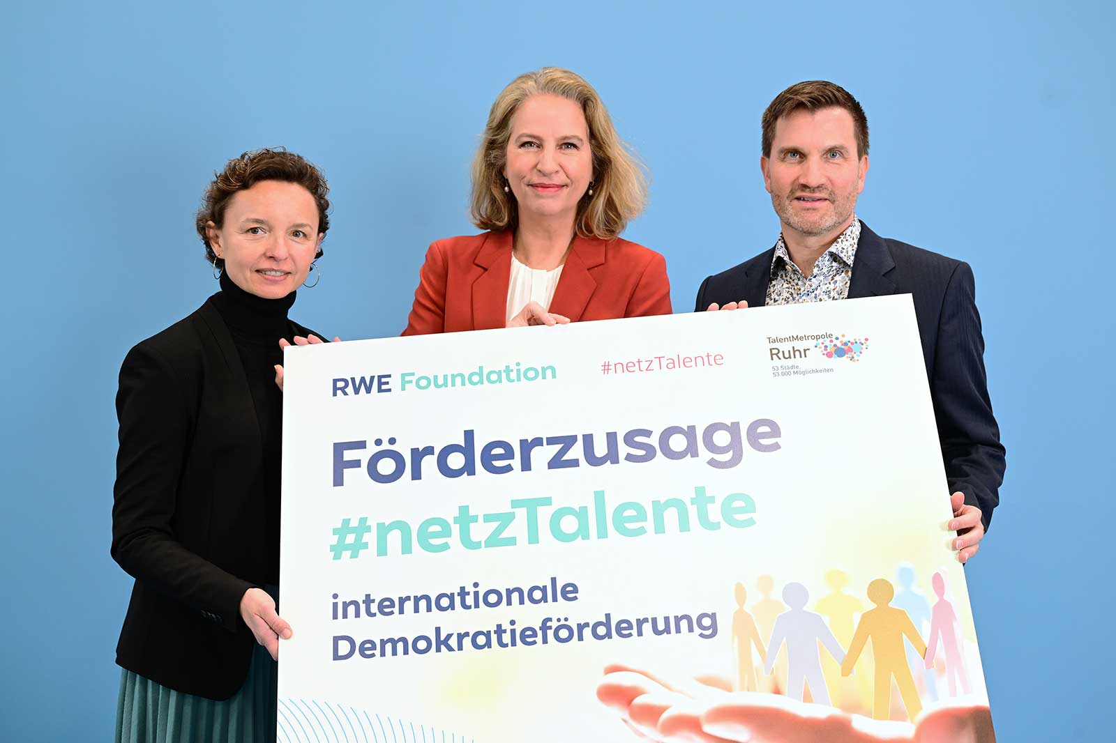 Three individuals are holding a large banner with the text 'Förderzusage #netzTalente internationale Demokratieförderung' against a blue background.
