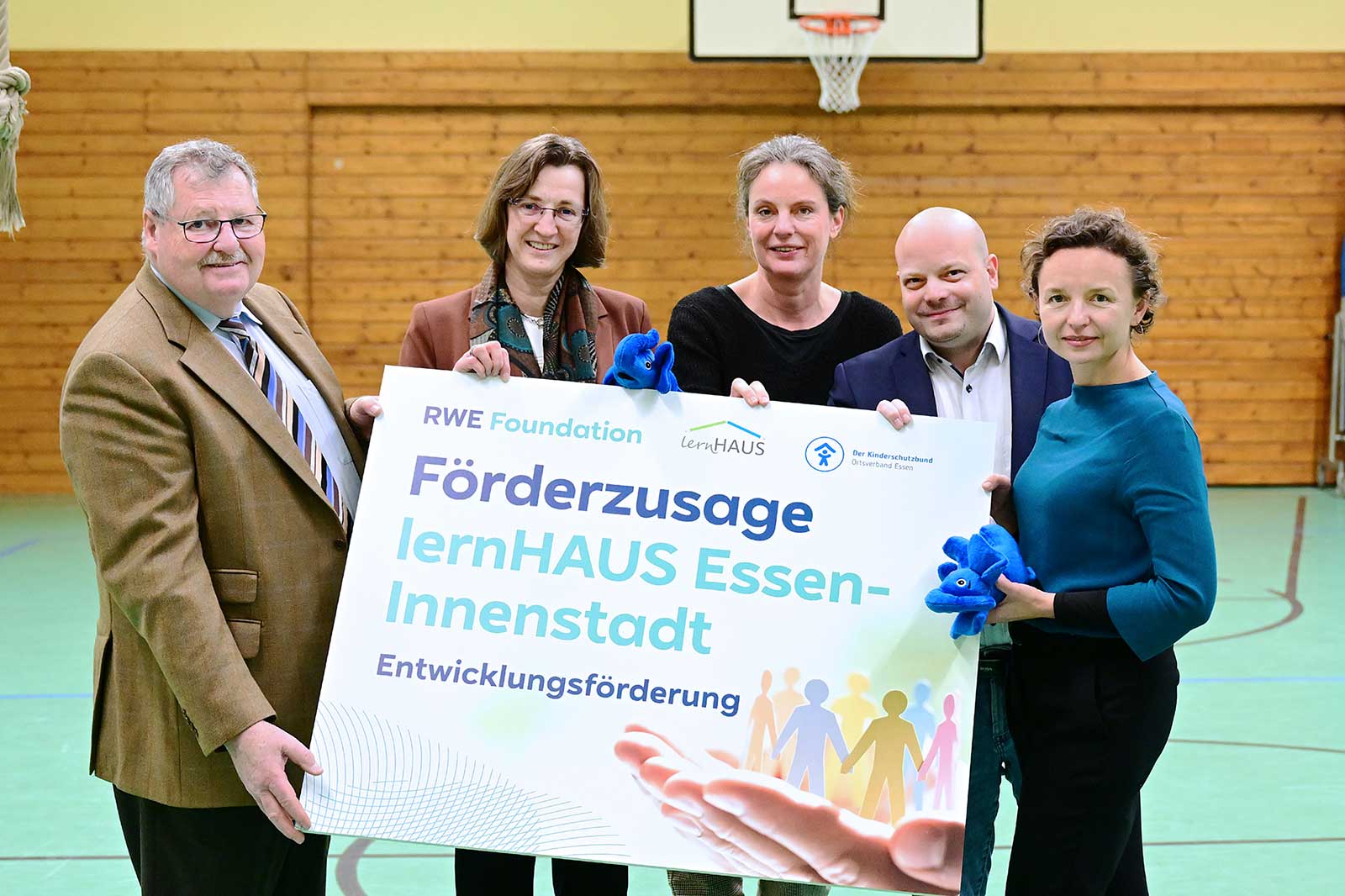 Five individuals hold a sign reading 'Förderzusage lernHAUS Essen-Innenstadt'. A basketball hoop is visible in the background.