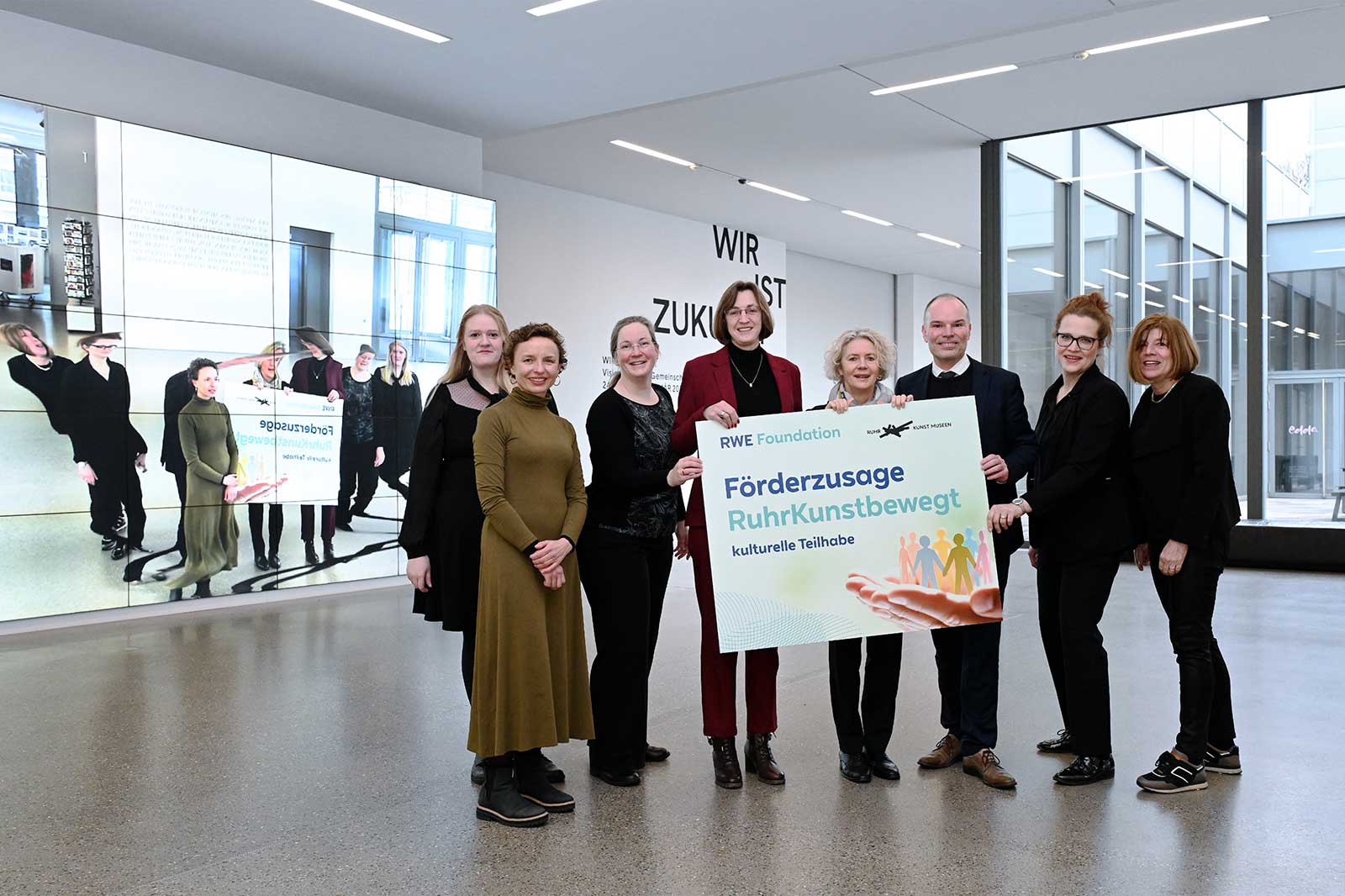 Eine Gruppe von Menschen hält ein Schild mit 'Förderzusage RuhrKunstbewgt' in einem modernen Raum.