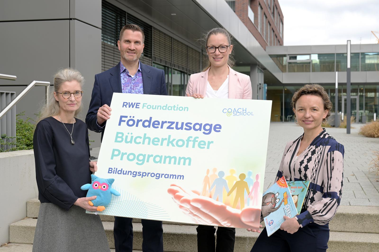 Vier Personen halten ein Schild mit 'Förderzusage Bücherkoffer Programm' im Freien. Die Umgebung ist modern und sauber.