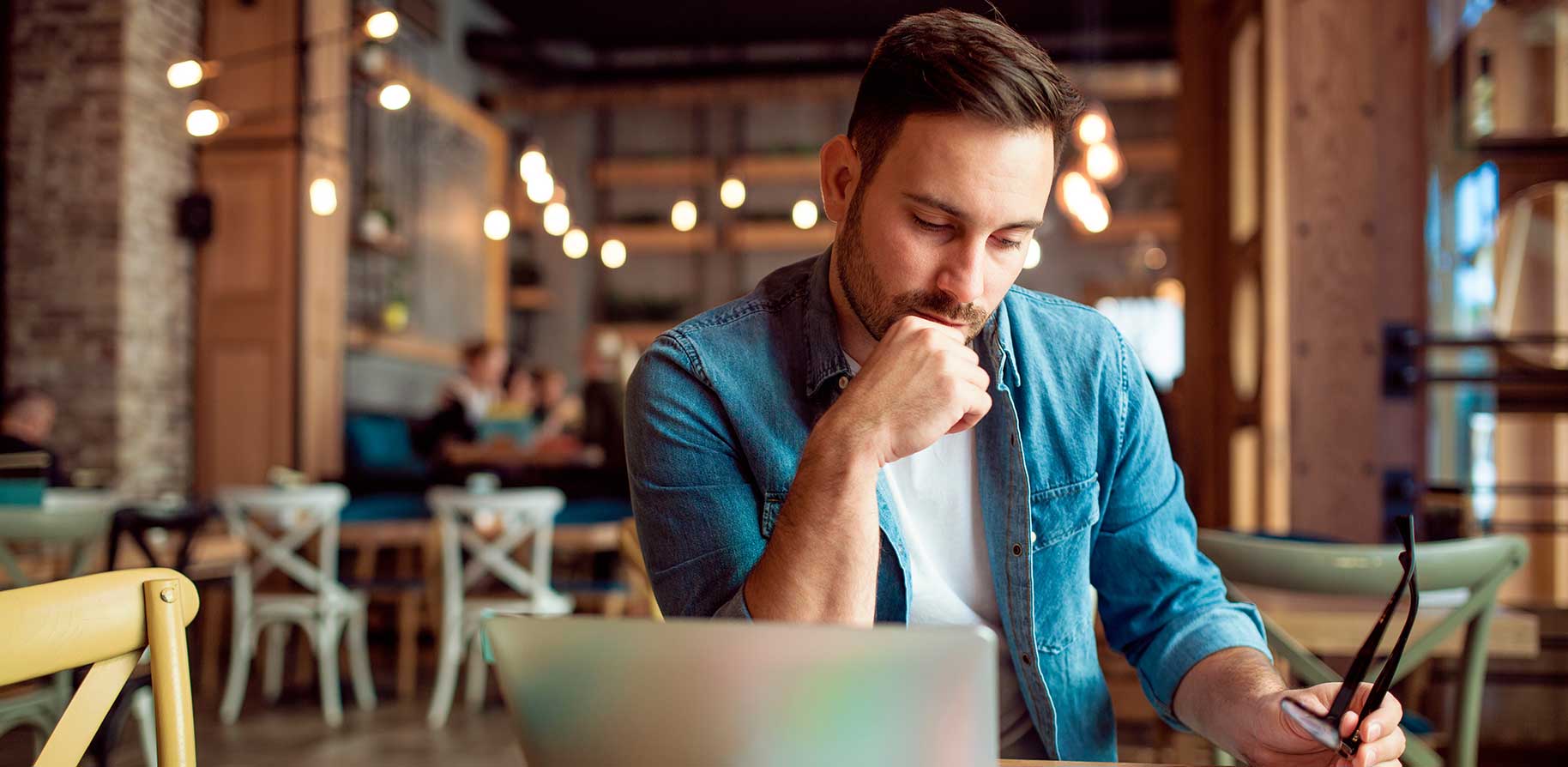 A man sits in a café at a table with a laptop, looking thoughtful. Chairs and tables are visible in the background.
