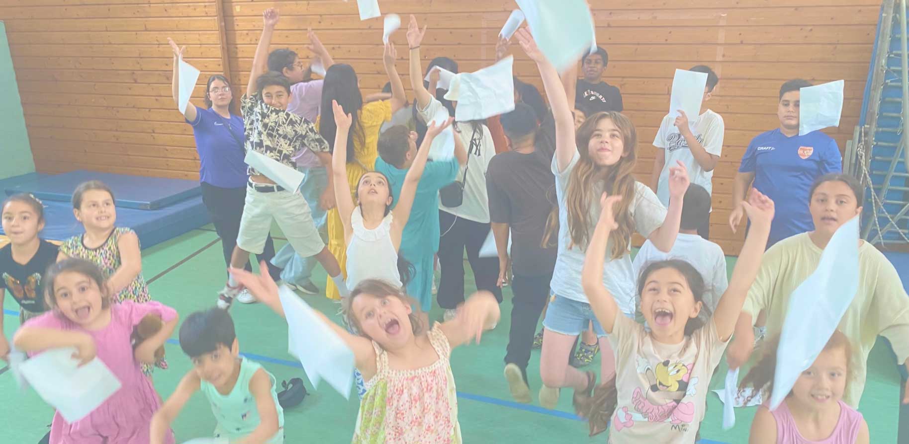 Eine lebhafte Gruppe von Kindern tanzt fröhlich und schwenkt Papiere in einer Turnhalle, die Energie und Begeisterung zeigt.